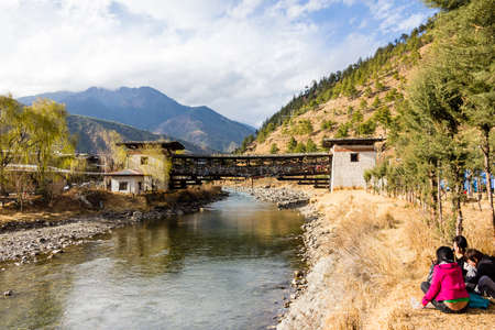 a typical bridge in Thimpu, Bhutan, covered with prayer flags  Three young women are sitting at the side of the river のeditorial素材