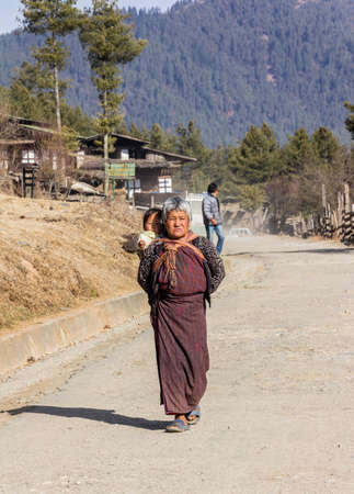 A woman in traditional bhutanese costume with a baby is walking through a village in the beautiful Phobjika valley, Bhutanのeditorial素材