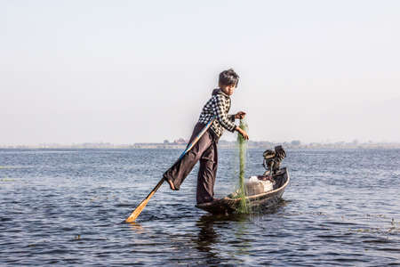 A legrowing fisherman at Inle lake is catching fish with a net . In the background is a village.のeditorial素材