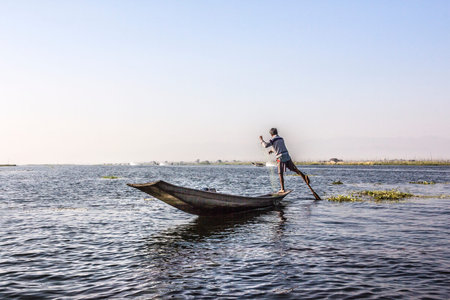 A legrowing fisherman at Inle lake is catching fish with a net . In the background are stilthouses floating gardens and the mountains.のeditorial素材