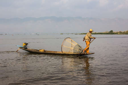 A fisherman at lake Inle, Myanmar stands in his boat, his fish trap is behind him.のeditorial素材
