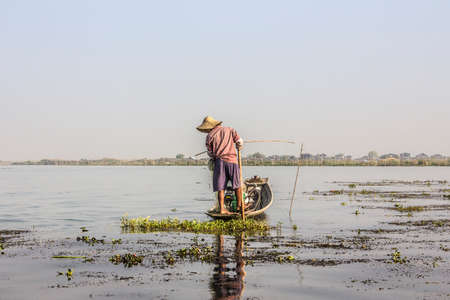 A fisherman at lake Inle, Myanmar stands in his boat and handles the net. In the background are the swimming gardens and stilt houses.のeditorial素材