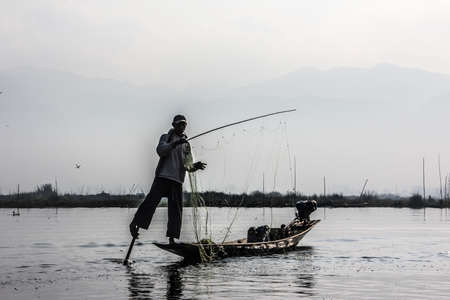 A fisherman at lake Inle, Myanmar stands in his boat and handles the net. In the background are the swimming gardens and stilt houses.のeditorial素材