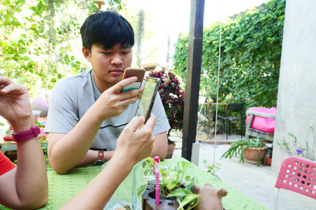 Asian teenage men smiling and looking at smartphone in his hand, People on Modern Technology at the coffee shop's dining table with green garden in background, Thailandの写真素材