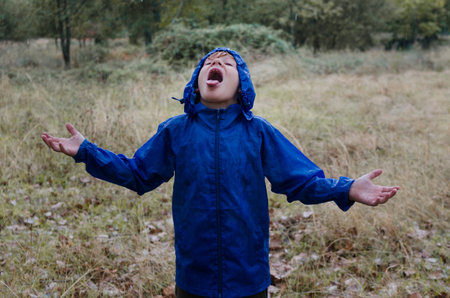 A 10 year old boy wearing a blue raincoat under the rain in the middle of nature with his mouth open...