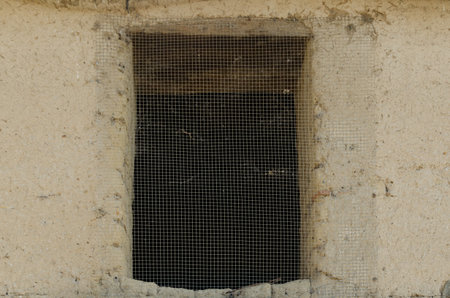 Front view of antique old adobe and stone wall with rectangular window hole covered with a metal...