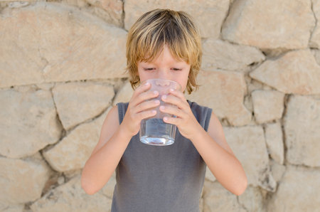 Cute blond caucasian little kid holding and drinking a glass of pure mineral water outdoors in summer. Healthy lifestyle and refreshment concept. Happy childhood. Copy spaceの写真素材