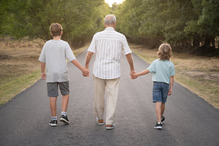 Horizontal photography of caucasian grandfather with his two grandchildren walking along a road in the middle of a forest in nature. Active and healthy retirement, family love and attachment conceptの写真素材