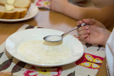 Close-up of a boys hand with a spoon of porridge over a plate. A boy is having breakfast at the dinner table. A plate of porridge is on a napkin.の写真素材