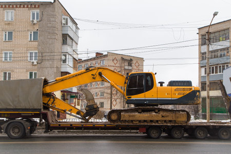 A heavy modern excavator stands on a low trailer platform along the rear ramp. Instead of a bucket,...