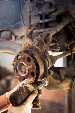 Male hands in gloves use a power tool to clean rust on the wheel hub of a car. In the garage, a man...