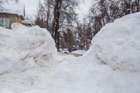 A wide large snowdrift by the road against the backdrop of a city street. On the road lies dirty snow in high heaps. Urban winter landscape. Cloudy winter day, soft light.の写真素材
