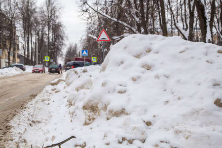 A large roadside snowdrift by the road against the backdrop of a city street. On the road lies dirty snow in high heaps. Urban winter landscape. Cloudy winter day, soft light.の写真素材