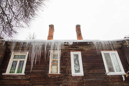 Spring transparent icicles hang on the edge of the roof. Against the background of the wooden wall of the old house. Large cascades, even beautiful rows. Cloudy winter day, soft light.の写真素材