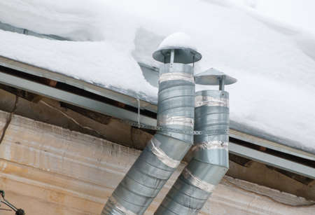 Two metal exhaust pipes on the roof of the house. There is white snow on the surface. Against the background of a gray sky. Cloudy winter day, soft light.の写真素材