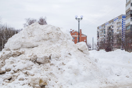 A powerful snowdrift by the road against the backdrop of city houses and trees. On the road lies dirty snow in high heaps. Urban winter landscape. Cloudy winter day, soft light.の写真素材
