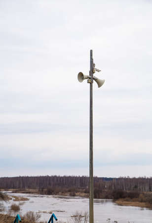 Two loudspeakers-megaphones on a high pole against the background of the river. Spring, the snow is melting, there are puddles of slush and mud all around. Day, cloudy weather, soft warm light.の写真素材