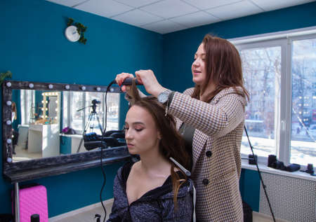 A young woman master with a smile twists a strand of the clients hair on tongs. The hairdresser makes a hairstyle for a young woman. Barber shop, business concept. Beauty salon, hair care.の写真素材