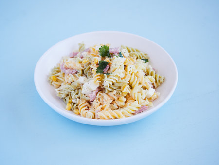 pasta salad plate with corn, cheese and jam, parsley on top, horizontal view on a blue surface with copy spaceの写真素材
