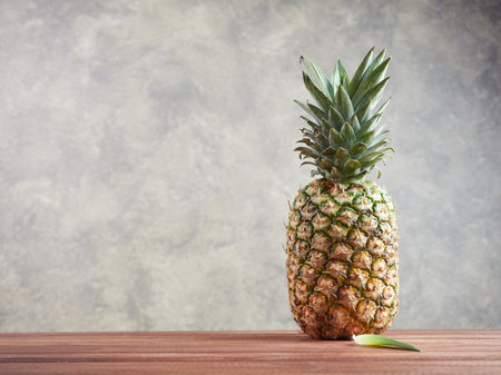 whole piece of fresh ripe pineapple on a wooden surface with a fallen leaf on a wooden surface, gray backgroundの写真素材