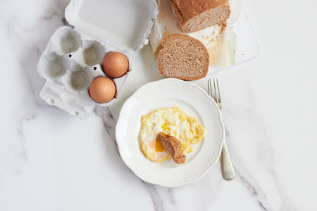 fried egg on a plate on a table with pieces of bread to dip in the yolk on a marble table with a fork beside it, slices of bread and two raw eggs in an egg cupの写真素材
