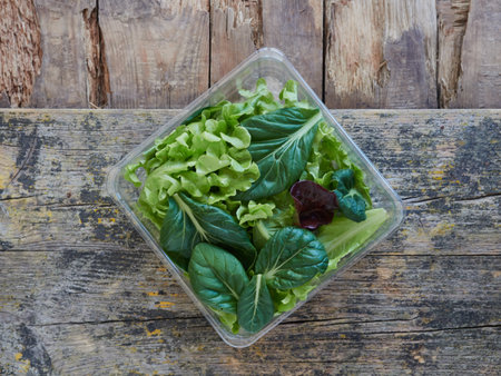assorted lettuce and vegetable leaves on a salad to take away on a square plastic packaging on a wooden surface, top viewの写真素材