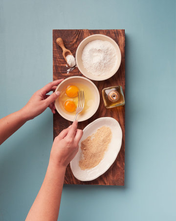 womans hands holding a fork to beat eggs with various cooking ingredients on a wooden board placed on a rectangle dark wooden board on a bluish background. View from aboveの写真素材