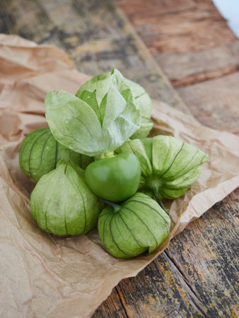 a pile of green tomatillo fruits on wrapping paper on a rustic wooden table. the top one is peeled and the others with the skinの写真素材