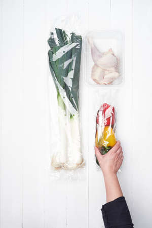 hand picking up colored peppers wrapped in plastic next to other plastic-wrapped food, leeks and chicken.concept of environmental problemの写真素材