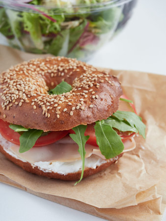 bagel filled with rocket, turkey ham, cream cheese and tomato on wrapping paper, take away salad on partial view blured backgroundの写真素材