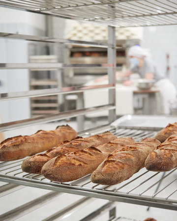 freshly baked baguette of bread on a cooling rack in interior of a bakehouse woman baker working in the background out of focusの写真素材