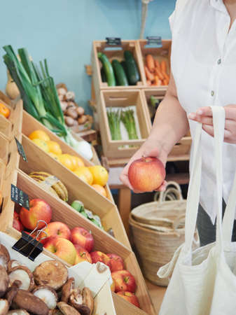 Partial view of an unrecognizable customer choosing an apple in a grocery store with a reusable cloth bag. Healthy and sustainable shopping concept in a small local store.の写真素材