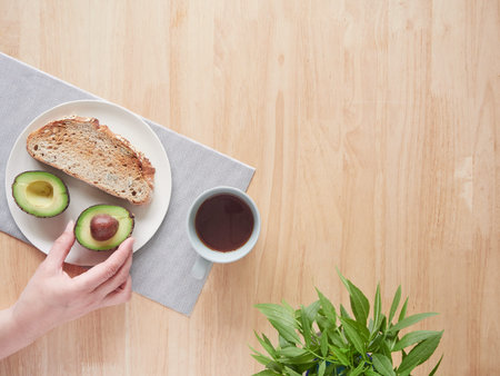 Woman's hand holding half avocado on a plate with toast at breakfast table. Top view with copy...