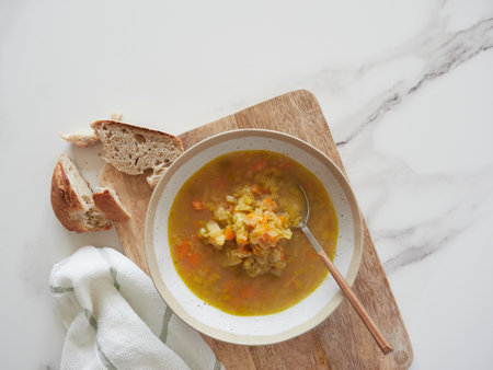 Top view of plate with vegetable soup and spoon on wooden cutting board and marble kitchen countertop. Portion of bread, crumbs and napkin.の写真素材