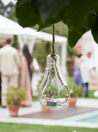 Outdoor wedding decoration: glass bulb hanging from tree with candle inside. Unrecognizable people...