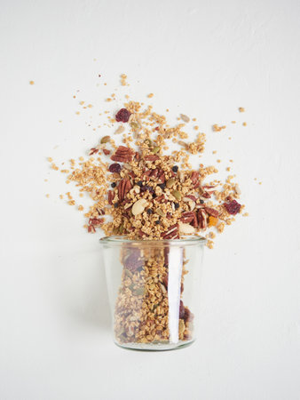 From above crispy granola with assortment of nuts scattered from glass on white tableの写真素材