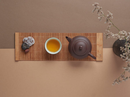 Top view of mug with hot tea placed between ceramic teapot and stone on wooden board near vase with...
