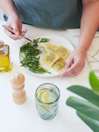 Partial view of adult woman at table with fork in hand, taking pasta (ravioli) from plate to eat at home. Side of salad of arugula greens, olive oil. Wooden pepper shaker and glass of water with lemon on white table.の写真素材