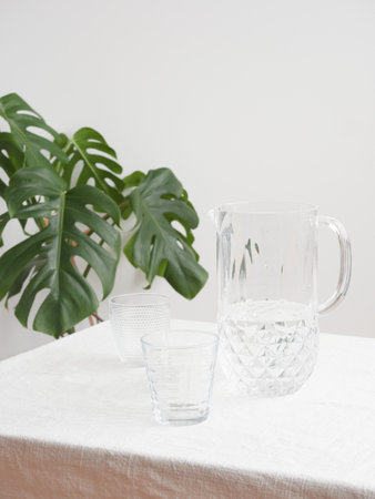 jar with fresh water and crystal glasses on table with tablecloth, green plant in the background with copy spaceの写真素材