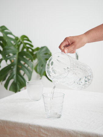 Drink water, healthy lifestyle. Concept of thirst and hydration. Woman's hand pouring fresh and pure water from a pitcher into a glass at the table.の写真素材