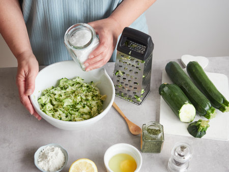 woman's hand with glass container of salt in her hand to season grated zucchini. On the countertop, other ingredients and utensils to prepare the recipe and cook.の写真素材