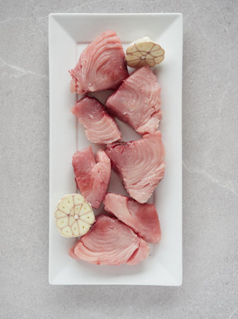 Fresh chunks of raw white tuna (Thunnus alalunga) called "bonito del norte" in Spain. On white rectangular plate with halved head of garlic. Top view on gray marble countertop background.の写真素材