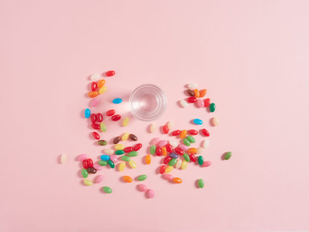 View from above glass of water and jelly beans as colored pills scattered on pink background. Concept of vitamins and food supplement.の写真素材