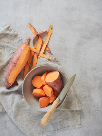 Top view of recipe preparation with sweet potato, peeled and sliced in gray bowl on kitchen towel next to knife.の写真素材