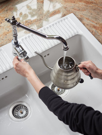 top view woman hands filling a kettle with tap water over the white kitchen sink. Tea or infusion preparation.の写真素材