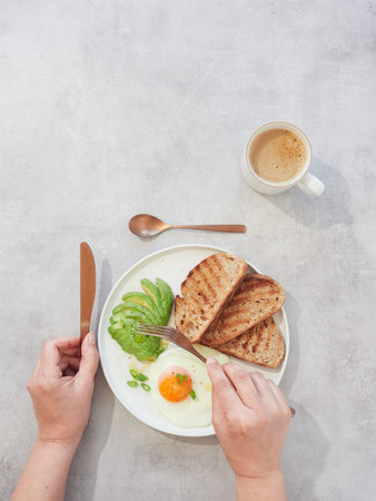Top view of woman hands eating breakfast with fried egg, bread and coffee on table