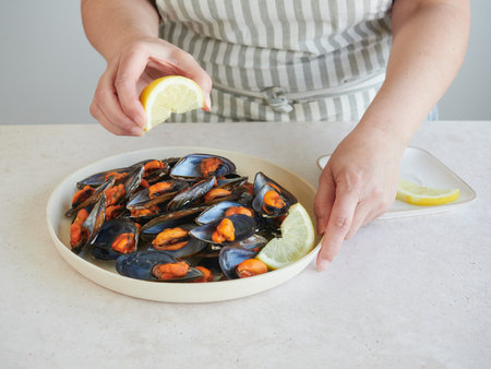 woman holding a lemon wedge in her hand over a plate of cooked mussels on the table.の写真素材