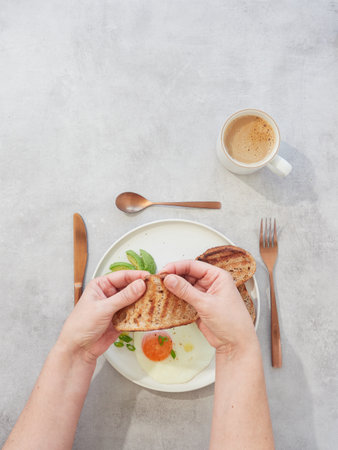 Top view of female hands holding toasted bread on plate with fried egg and avocado. Coffee in cup for breakfast or brunch.の写真素材