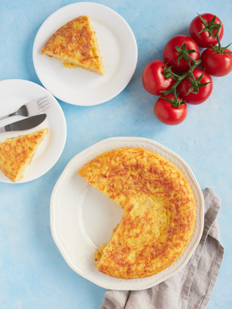 Typical homemade Spanish potato omelette, triangular portions served on plates. Fresh red tomatoes on blue background.の写真素材