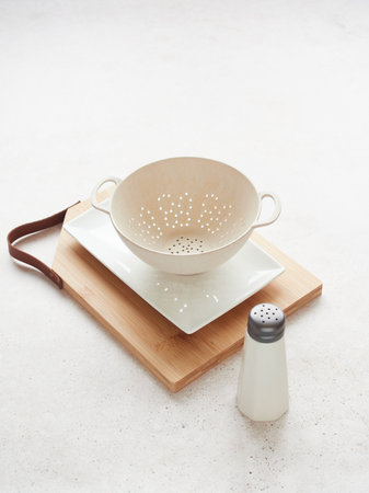 Small empty colander on white plate and wooden cutting board, next to salt shaker on the table.の写真素材